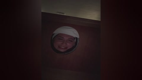 A dimly lit video shows a person smiling from inside a wooden cabinet. The person is wearing a white helmet, their face framed by a circular opening. The background is dark and shadowy.
