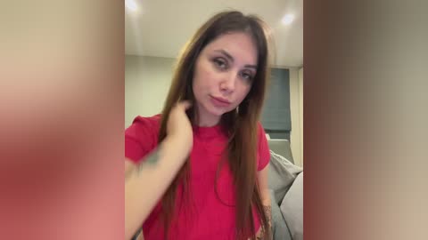 Video of a young Caucasian woman with long, straight brown hair, wearing a red t-shirt, standing in a modern, beige-toned room with a grey couch and a white fridge in the background.