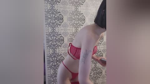 Video of a slender, light-skinned woman with a short black bob haircut, wearing red lace lingerie. She stands in front of a gray and white damask-patterned shower curtain, holding a smartphone.