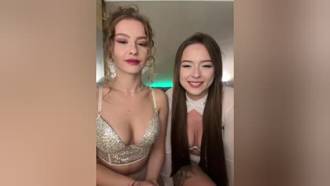 Video of two young women with light skin, one with curly blonde hair, the other with long brown hair, both smiling, wearing revealing outfits in a dimly lit room.