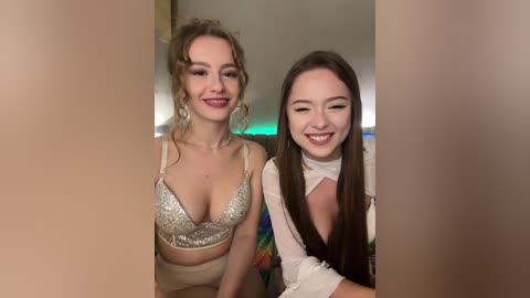 Video of two women smiling; one with curly hair in a glittery bra, the other with long hair in a white top, in a dimly lit room.