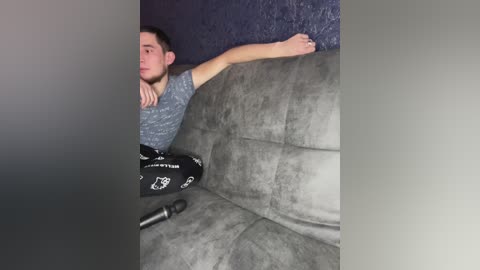 A video of a young man with short dark hair and a beard, wearing a gray t-shirt and black pants, lying on a gray suede couch against a dark blue wall.