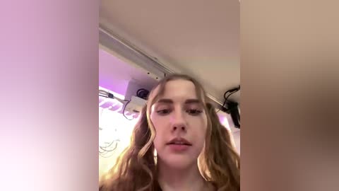 A young woman with wavy, shoulder-length blonde hair, fair skin, and full lips is seen from the waist up, looking slightly to the left. The background features a ceiling with recessed lighting and a vent.