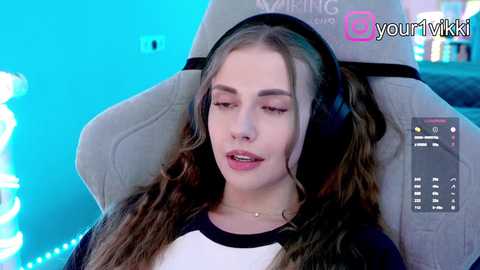 Video of a young woman with long brown hair, wearing headphones, sitting in a grey gaming chair. She has fair skin and a serene expression. Background features a blue, lit-up room with a gaming controller.