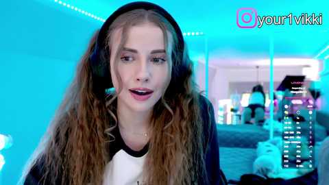 Video of a young woman with long, wavy brown hair, wearing headphones, smiling, sitting in a brightly lit, modern room with a TV screen and gaming console in the background.