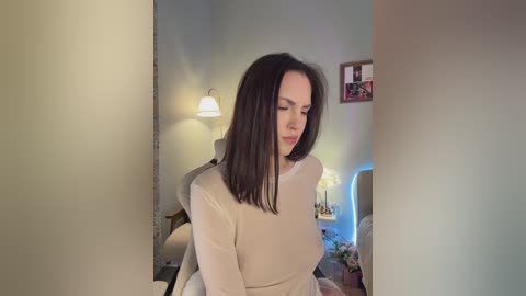 Video of a fair-skinned woman with straight, shoulder-length dark brown hair, wearing a beige long-sleeved top, sitting on a beige chair in a softly lit, cozy room with a white lamp, framed photos, and a blue chair in the background.