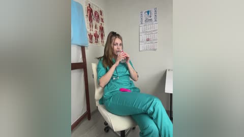 Video of a young Caucasian woman with long brown hair, wearing teal scrubs, drinking from a pink cup while sitting on a white medical chair in a sterile exam room with anatomical posters and a calendar on the wall.