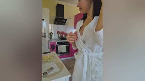 Video of a slender woman with long black hair in a white terrycloth robe, standing in a modern kitchen with pink cabinets and stainless steel appliances.