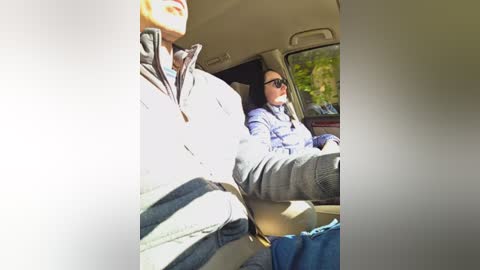 A video shows a person in sunglasses, wearing a grey jacket and jeans, sitting in a car. The interior is bright, with sunlight streaming through the window, highlighting a green landscape outside.