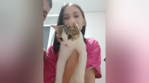 A video of a young woman with long black hair, wearing a pink shirt, holding a white and brown tabby cat against a blurred, pink wall background.