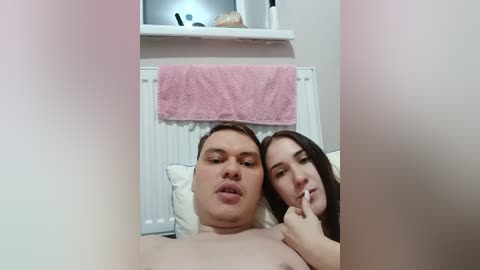 Video of a nude, overweight man with light skin and a chubby face, lying on a bed. A slim woman with long brown hair, also nude, is partially covering his face with her hand. Pink towel hangs on a radiator.