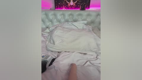 Video of a messy bed with white pillows and sheets, a remote control on the left, a hand reaching in, and a wall with a colorful, abstract painting in the background.