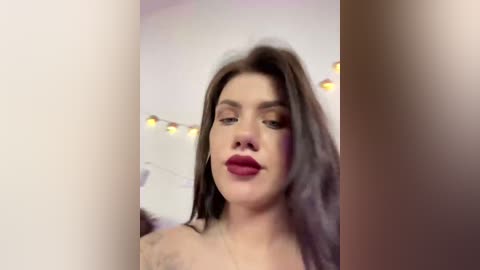 Video of a young woman with light skin and long brown hair, wearing bold red lipstick, in a softly lit room with warm, fuzzy lighting and white walls.