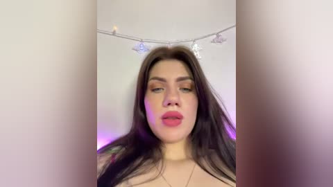 A video of a fair-skinned, long-haired woman with dark eyebrows, red lipstick, and subtle makeup, wearing a necklace, against a softly lit background with fairy lights.