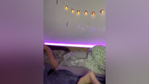 Video of a messy bedroom with a person in a floral-patterned bed, surrounded by a string of fairy lights and a purple LED strip on the wall.