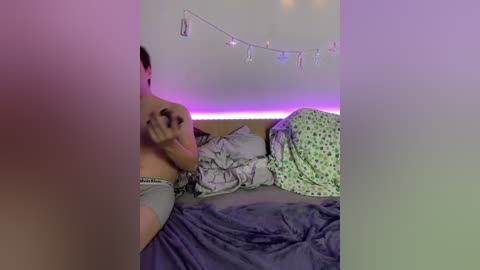 Video of a young woman in a bedroom with purple LED strip lighting, wearing grey boxer shorts, and a messy bed with green and grey patterned sheets.