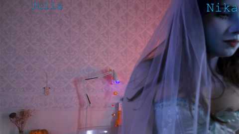 A video featuring a young woman with fair skin and long, straight brown hair, wearing a sheer, light-colored dress. The background shows a white wall with intricate floral wallpaper, and a small, ornate table with a glass vase. The image is dimly lit with a pinkish hue.