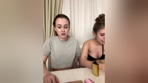 Video of two women with fair skin and light brown hair, one in a gray crop top and the other in a black bra. They sit at a table, one applying makeup, while the other watches. Background features beige curtains and a light-colored wall.