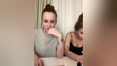 A video of two women, one with a bun and the other with brown hair, sitting at a table, wearing casual clothing, looking introspective.