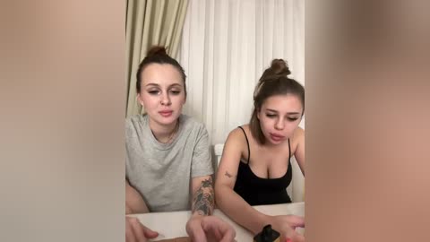 Video of two women sitting side by side at a table, one with light skin and brown hair in a bun, wearing a gray shirt; the other with light skin, dark hair in a bun, and a black top. Background features a beige curtain.