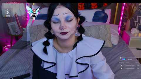 Video of a woman with white face paint, blue eyeshadow, and black hair in pigtails, wearing a white ruffled blouse, lying on a bed with a gray and white quilt.