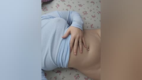 A video showing a light-skinned person lying on a floral-patterned bedspread, wearing a light blue long-sleeve shirt and no pants, hand resting on their stomach.