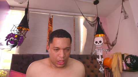 A video shows a shirtless man with short black hair and medium skin tone, wearing a confused expression. The background features Halloween decorations, including a skeleton, a witch, and a bat hanging from the ceiling. The room has a dark gray upholstered headboard and orange pumpkins.