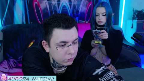 Video of two young people in a dark, neon-lit room; a guy with black hair, tattoos, and a chain necklace, and a girl with blue hair and a black top.