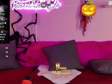 Video of a vibrant Halloween-themed room with a glowing \"Boo\" sign, pumpkin lights, and a red couch adorned with black spider webs, surrounded by plush pillows. A \"Happy Halloween\" banner and a jack-o'-lantern are prominent.