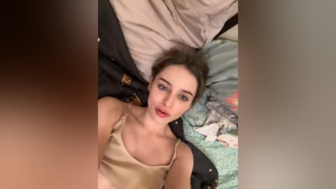 A video of a young woman with fair skin, brown hair, and full lips, lying on a bed with messy pillows and clothes, wearing a beige camisole.