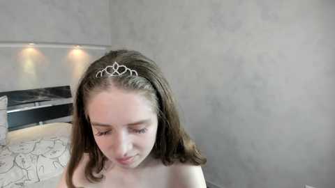 A young Caucasian woman with long brown hair, wearing a silver tiara, sits on a bed with white sheets and a black headboard in a modern, minimalist bedroom.