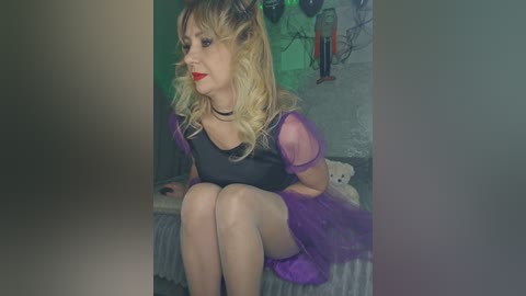 Video of a blonde woman with wavy hair, wearing a sheer purple dress and black choker, sitting on a bed, surrounded by a dimly lit room with green lighting and a teddy bear.