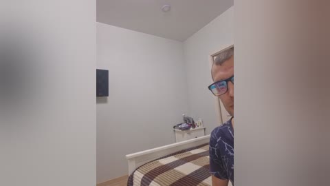 A video of a small, sparsely decorated bedroom with a man in glasses peering through a doorway, wearing a navy shirt. The room features a white bed with a striped blanket, a small desk, and a dark wall-mounted TV.