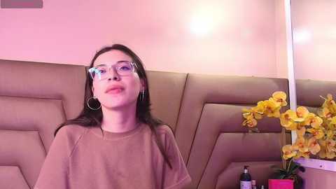Video of a young woman with straight black hair, wearing glasses, a beige t-shirt, and hoop earrings, sitting on a beige padded bench, with a vase of yellow orchids and a bottle of sanitizer in the background.
