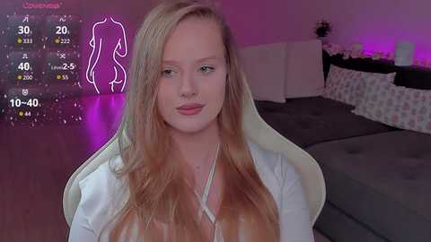 Video of a fair-skinned woman with long, straight blonde hair, wearing a white top, sitting in a grey chair in a dimly-lit bedroom. A digital display shows her stats and a neon outline of a woman in the background.