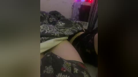A blurry, grainy video shows a person lying on a bed with a floral-patterned duvet, revealing their lower abdomen and legs. The background features a dimly lit room with red and purple lighting.
