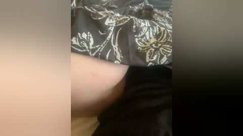 Video of a person's arm and upper chest, wearing a black shirt with gold floral designs. The skin tone is fair, and the texture of the shirt appears smooth. The background is blurred.