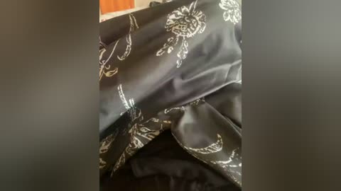 A video of a black satin garment with intricate white floral embroidery, draped over a surface, revealing a soft, glossy texture. The background is blurred, emphasizing the fabric details.