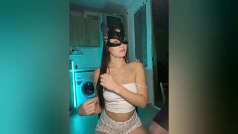 Video of a young woman in a black leather mask, white crop top, and patterned shorts, kneeling on a light-colored floor, in a dimly lit room with a washing machine and kitchen cabinets.