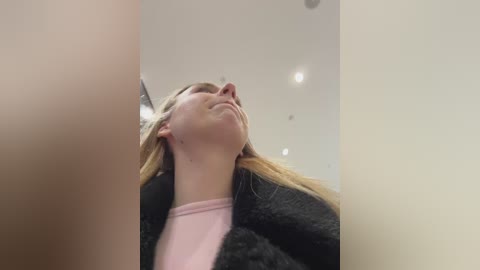 A video of a young Caucasian woman with blonde hair, wearing a pink shirt and a black fur coat, looking up with a serene expression.