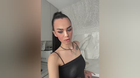 Video of a slender woman with fair skin, wearing a black, shiny dress, and a necklace. She has a high ponytail, sharp eyebrows, and dramatic makeup. Background features a white bed and a white textured wall.