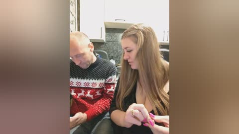 Video of a bald, middle-aged man and a young woman with long brown hair in a black sweater, sitting closely together in a modern kitchen.