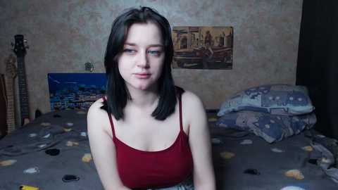 Video of a young woman with straight black hair, wearing a red spaghetti-strap tank top, sitting on a bed with patterned bedding in a dimly lit room with posters on the wall.