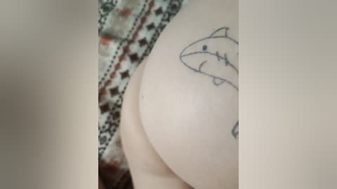 Video of a person's bare upper torso, with a prominent tattoo of a fish on the right side of their chest. The skin is light, and the background shows a patterned fabric.