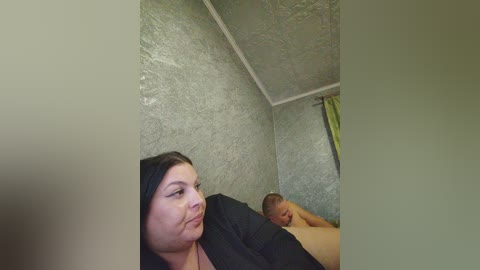 Video of a smiling woman with light skin and dark hair, lying on a bed with a man who has a shaved head, in a room with textured green walls and a yellow curtain.