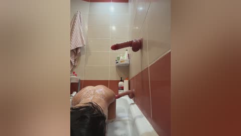 A video of a man with a large, erect penis standing in a small, tiled bathroom. The room has beige and brown walls, a red towel, and toiletries on a shelf.