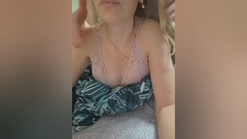 Video of a blonde woman with fair skin, wearing a pink lace bra and a green and white patterned blanket. She is in a bed, with a blurred male figure partially visible.