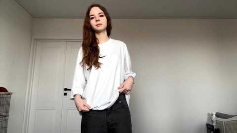 Video of a young, fair-skinned woman with long brown hair, wearing a white Nike sweatshirt and black pants, standing in a sparsely furnished, plain white room with a laundry basket and white door.