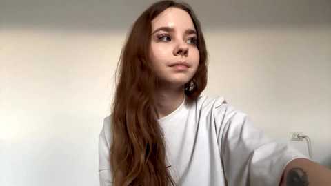 Video of a young girl with long, straight brown hair, wearing a white t-shirt, looking thoughtfully off-camera. Background is a plain, light-colored wall.