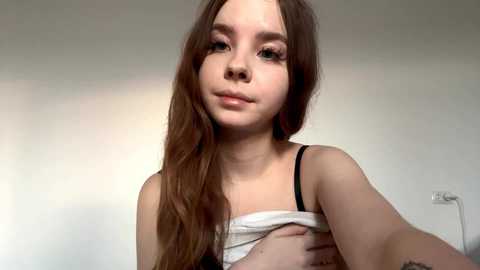 Video of a young Caucasian girl with long brown hair, fair skin, and brown eyes, wearing a white tank top. She has a neutral expression. Background is a plain, light-colored wall.